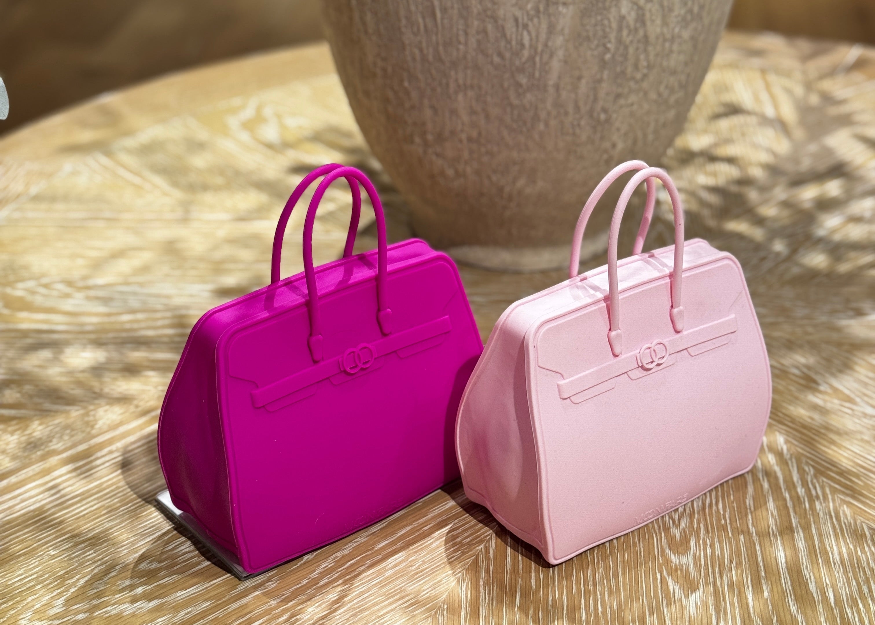 Two small pink and purple handbag-shaped objects on a wooden surface with a vase in the background.